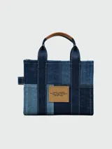 Cartera tipo tote de Marc Jacobs, confeccionada en denim con diseño de paneles en diferentes tonos de azul. Presenta el logo de la marca estampado en el frente, asas superiores de mano y una correa de hombro ajustable y extraíble en color azul oscuro.