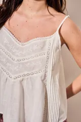 Blusa color off white de algodón peinado, sin mangas, con tirantes finos, escote en V con detalles de encaje y bordados, y pliegues decorativos en los laterales.