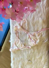 Conjunto de lencería compuesto por un bralette y una tanga, confeccionados en encaje bitono rosa y champagne con diseño floral. El bralette presenta cierre frontal con broche de gancho y breteles regulables. La tanga es de estilo colaless con laterales regulables.