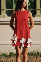 Vestido corto rojo sin mangas, con escote redondo y silueta recta. Presenta detalles de flores tridimensionales aplicadas en el borde inferior, con pétalos blancos y tiras colgantes.