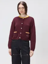 Cardigan de algodón grueso en color rojo, con cuello redondo y cierre frontal de botones grandes. Presenta bolsillos cuadrados en el bajo con detalles de pelo sintético en los bordes y logo bordado en uno de ellos. Posee puños y ruedo elastizados con diseño holgado.