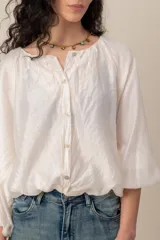 Blusa blanca de diseño amplio, con mangas abullonadas, escote redondo fruncido y cierre frontal de botones nacarados. Cuenta con elástico en la cintura que genera efecto blusón.