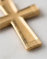 Dije en forma de cruz de acero inoxidable con acabado dorado y textura en los extremos.