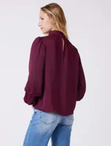 Blusa Tahari de gasa satén color violeta oscuro, con cuello alto fruncido y mangas largas abullonadas. La espalda tiene una abertura central con cierre de botón.