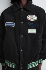 Cazadora estilo varsity confeccionada en pana de algodón azul, con interior ligeramente acolchado. Presenta cuello solapa, cierre frontal con botones a presión, bolsillos laterales de vivo y acabados en rib a rayas en puños y cintura. Incluye múltiples parches bordados decorativos en el frente.