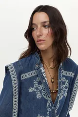 Camisa de denim azul con bordados blancos en el frente y las hombreras. Cuenta con cuello mao y cierre frontal con botones a presión.