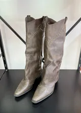 Botas texanas de caña alta color marrón con detalles de tachas doradas. El calzado presenta un diseño con puntera en punta y un pequeño taco cuadrado. Se recomienda pedir un talle más del que se usa habitualmente.