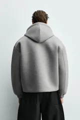 Sudadera gris de corte boxy fit con capucha y cierre frontal de cremallera. Confeccionada en tejido doble y compacto, cuenta con bolsillos laterales ocultos en la cadera y mangas largas con puños elásticos.