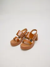Sandalias de cuero color marrón con plataforma y taco cuadrado. Presentan dos tiras anchas sobre el empeine y una tira trasera con elástico. Decoración de doble argolla en los laterales.