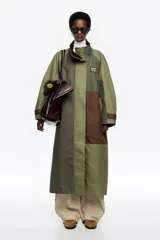 Gabardina larga de diseño patchwork en tonos verdes y marrones, con cuello alto, cierre frontal oculto y logo de la marca en el pecho. Presenta un corte amplio y un interior acolchado para mayor abrigo.