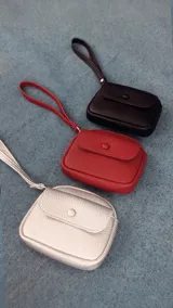 Monedero compacto de símil cuero con textura granulada, cuenta con una correa de mano, cierre principal y un bolsillo frontal con solapa y botón a presión.