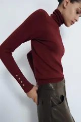 Jersey de punto liso en color beige, con cuello alto y manga larga con detalle de botones metálicos en los puños.