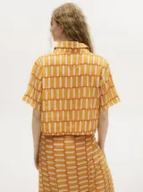 Camisa crop de manga corta con cuello camisero y cierre frontal de botones. Presenta un diseño geométrico estampado en tonos naranja y crema, con un calce suelto y liviano.