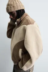 Cazadora de corte relajado confeccionada en tejido de borreguillo con diseño de paneles en tonos marrón y beige. Presenta cuello subido, cierre frontal de cremallera, bolsillos laterales y acabados en rib en puños y cintura.