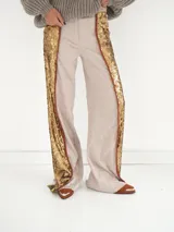 Pantalón acampanado de tiro medio, confeccionado en tela brocada con estampado floral en tonos beige y dorado, con franjas laterales de lentejuelas doradas y ribetes de cuero marrón.