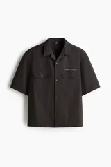 Camisa cargo de manga corta color marrón oscuro, con cuello abierto, botones frontales, bolsillos con solapa en el pecho y el texto 'SERVICE GARMENTS' bordado sobre el bolsillo izquierdo.