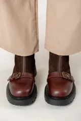 Bota de caña corta combinada en cuero liso y gamuza color marrón. Presenta cierre frontal con cremallera, tira con hebilla metálica decorativa en el empeine y suela track de goma negra.
