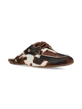 Zueco de cuero con pelo con estampado animal print tipo vaca en tonos marrones, blancos y negros. Presenta un diseño destalonado con cordones decorativos en la parte superior y ojales metálicos.