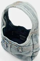 Bolso de mano tipo hobo confeccionado en tejido denim con diseño inspirado en un jean, incluyendo detalles de bolsillos exteriores, costuras reforzadas y trabillas. Cuenta con cierre de botón imantado y asa integrada.