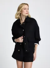 Chaqueta corta negra de tela tejida con cuello alto, mangas largas con puños abotonados, dos bolsillos delanteros con solapa y cierre frontal con botones.