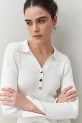 Sweater de punto acanalado color blanco, con cuello tipo polo y botonadura frontal de botones metálicos dorados. Tiene mangas largas con terminación en pequeñas aberturas.