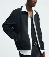 Campera masculina de corte boxy en color beige, con cuello inglés, cierre frontal de cremallera, mangas largas con puños elásticos y ruedo con elástico. Presenta bolsillos laterales y detalles de recortes en el diseño.