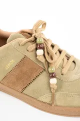 Zapatilla de gamuza en tono verde pistacho con detalles en cuero marrón, cordones al tono con cuentas decorativas y suela de goma color caramelo.