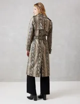 Trench de ecocuero con estampado animal print de serpiente en tonos beige, marrón y negro. Presenta cuello con solapa ancha, doble abotonadura frontal con botones negros, cinturón ajustable con hebilla metálica, mangas largas con presillas y botones en los puños, canesú suelto en la espalda, bolsillos laterales y forro interno.
