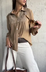 Camisa de color camel con cuello clásico, manga larga y detalle de bordado geométrico en color blanco en la parte frontal superior. Presenta cierre frontal con botones y un corte holgado.