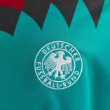 Camiseta de fútbol Adidas retro de la selección de Alemania, diseño alternativo de 1994. Color principal verde turquesa con cuello en V amarillo, rojo y negro, y un llamativo estampado geométrico en el pecho y las mangas en tonos negro, rojo y amarillo.