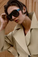 Trench corto de color beige, con cuello de solapa, manga larga con hombreras y trabillas con botones. Cuenta con cierre frontal cruzado con botones oscuros y cinturón ajustable del mismo tejido.