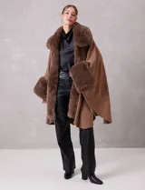 Ruana de tejido color tostado con cuello, bordes frontales y puños amplios con apliques de piel sintética al tono. Presenta una abertura central con cierre de botón tipo trenca.