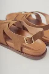 Sandalias planas de gamuza color marrón, con diseño de tiras cruzadas en el empeine y cierre con hebilla metálica dorada en el tobillo. Cuenta con plantilla de cuero y suela de goma con textura.