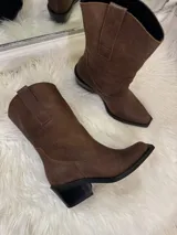 Bota estilo texana de caña media, confeccionada en gamuza color marrón. Presenta puntera ligeramente cuadrada, taco cuadrado de altura media en color negro y costuras reforzadas en el diseño de la caña.