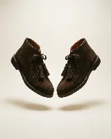 Bota de caña media confeccionada en cuero gamuzado color marrón chocolate. Presenta un diseño de estilo mocasín con cordones frontales decorados con borlas desmontables y suela plana de goma resistente.