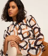 Blusa de chiffon con estampa de eslabones y argollas entrelazadas en tonos marrones y negros sobre fondo blanco. Presenta cuello alto, mangas largas acampanadas y cierre tipo gota en la espalda.