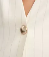Chaleco sastre sin mangas, color blanco hueso con finas rayas verticales en tono más claro. Presenta un profundo escote en V con un solo botón dorado central y bolsillos falsos con solapa en la parte inferior delantera.