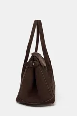 Bolso tipo citybag confeccionado en serraje color marrón oscuro. Presenta una solapa frontal con cierre magnético, asas de hombro integradas y detalles de hebillas metálicas en los laterales para ajuste. Incluye bolsillo interior con cremallera.