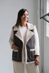 Chaleco de gamuza sintética en color gris oscuro con interior y bordes de corderito beige. Presenta solapas amplias, dos bolsillos frontales con ribetes de corderito y diseño sin cierre.