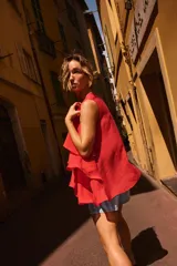 Top de cuello redondo y manga sisa, confeccionado en tejido con acabado satinado. Presenta volantes en cascada y un diseño de bajo asimétrico, con cierre en la espalda mediante pequeña abertura y botón.