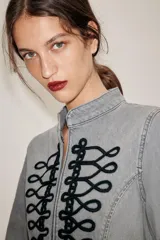 Chaqueta de denim gris con cuello subido y manga larga. Presenta un diseño entallado con bajo en evasé y un llamativo bordado frontal de alamares en color negro. Cierre frontal con corchetes ocultos.