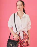 Cartera negra de cuero charolado con solapa y cierre de broche. Presenta un detalle metálico dorado en forma de freno de caballo en la parte frontal y una correa ajustable.