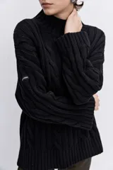 Sweater de punto con diseño de trenzas, cuello alto y mangas largas con puños acampanados. Presenta un corte holgado con terminación asimétrica en el ruedo.