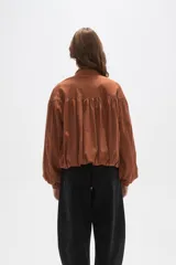 Chaqueta tipo bomber color tostado, confeccionada en material efecto gamuza. Presenta diseño con frunces en el pecho y espalda, cuello mao, cierre frontal completo y mangas largas con puños elásticos.