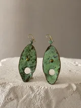 Aros colgantes de diseño orgánico con forma de hoja, elaborados en cobre con acabado de pátina verde oxidada y textura irregular. Presentan un detalle de remache central, un calado circular en la parte inferior y un pequeño dije de piedra turquesa en la parte superior, con pasante de plata.