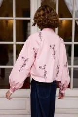Camisa de algodón color blanco con cuello mao y cierre frontal de botones. Presenta un diseño de bordados florales en color negro distribuidos en el frente y las mangas. Posee mangas largas abullonadas con puños abotonados y un corte recto ligeramente cropped.