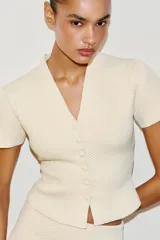 Chaqueta de punto color crudo con cuello en pico y manga corta abullonada. Presenta cierre frontal con botones a tono y una textura tejida estructurada.