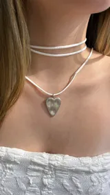 Gargantilla tipo choker compuesta por una tira de gamuza blanca que rodea el cuello y un dije de corazón metálico plateado con acabado texturizado.