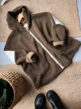 Campera tipo buzo con capucha confeccionada en tela sherpa, con diseño artesanal en la espalda que representa un paisaje de campo con sol y luna mediante apliques de tela. Presenta una combinación de colores verde oscuro y crudo, con cierre frontal y corte holgado.