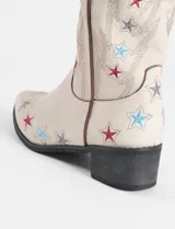 Botas estilo tejano de caña alta, confeccionadas en cuero vegano color beige. Presentan un diseño bordado con estrellas en tonos rojo, azul y gris, con taco cuadrado bajo y punta ligeramente afinada.